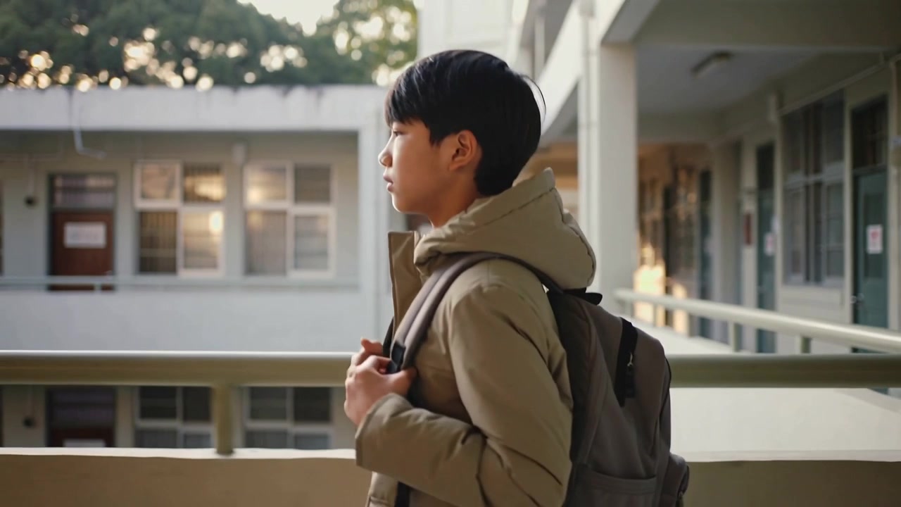 학교 난간에 다다른 카키색 코트를 입은 십대가 회색 배낭을 들고 잠시 멈춰 서서 안뜰을 바라보는 모습