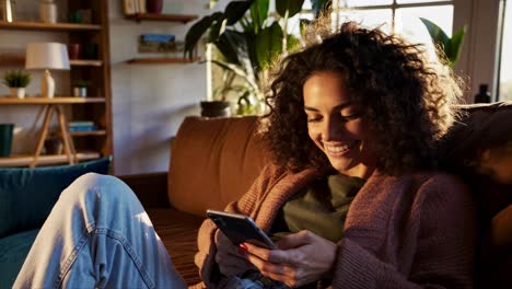 A woman with curly hair sits on a couch, smiling at her phone. The video captures a warm, relaxed