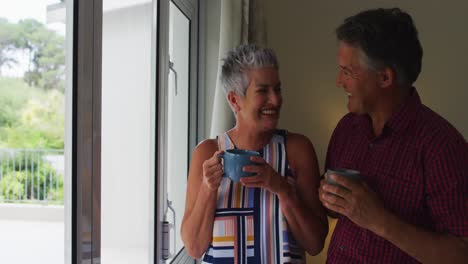 Kaukasisches-älteres-Paar-Lächelt-Und-Trinkt-Gemeinsam-Kaffee,-Während-Es-Zu-Hause-Aus-Dem-Fenster-Schaut