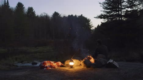 Man-sitting-by-a-campfire-with-a-lantern-and-blanket-in-a-forest-at-dusk,-surrounded-by-trees-and-nature