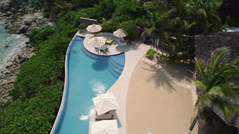 Aerial-over-luxurious-tourist-villa-with-outdoors-swimming-pool-framed-by-swaying-palm-trees-in-Huatulco,-Mexico