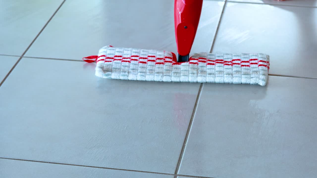 A red and white spray mop glides across a clean white tile floor in a sunlit indoor space, demonstrating efficient modern cleaning technique with smooth camera movement
