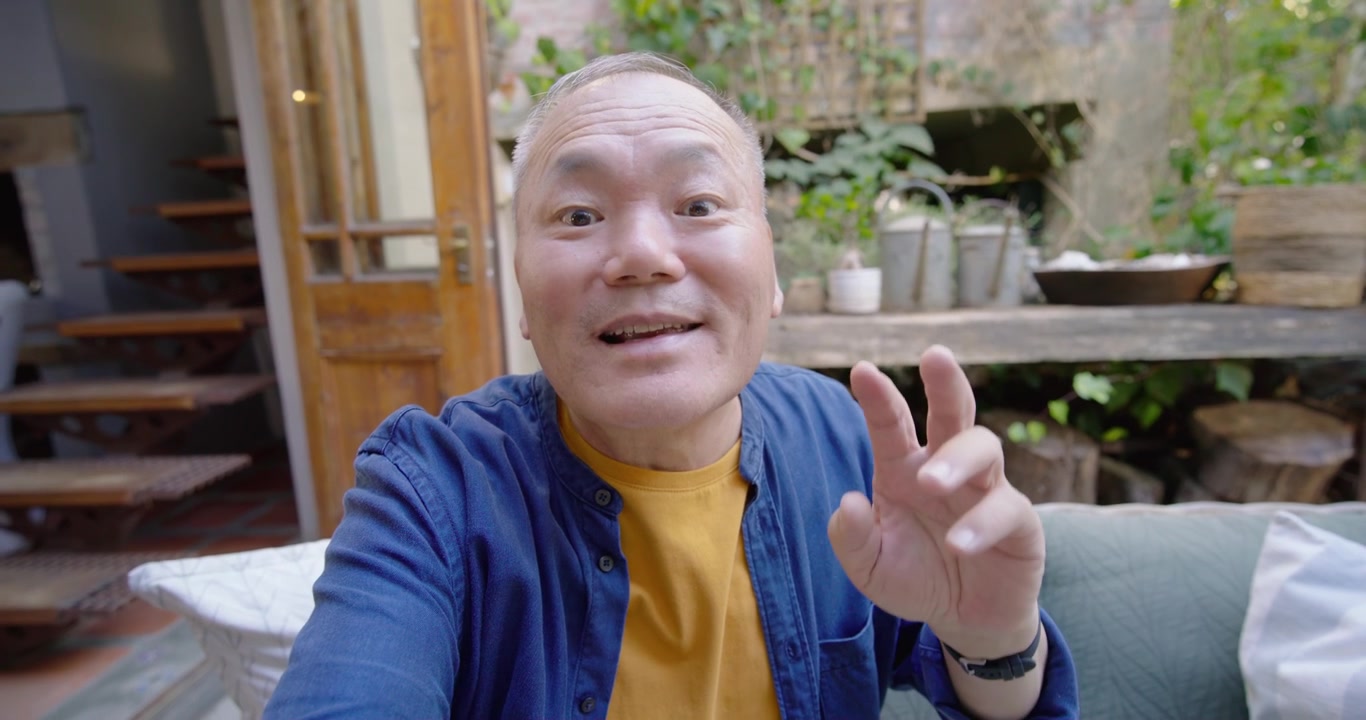 Anziano uomo asiatico che tiene una fotocamera per selfie, registra un saluto, sorride, indica e saluta con la mano sul divano del patio.