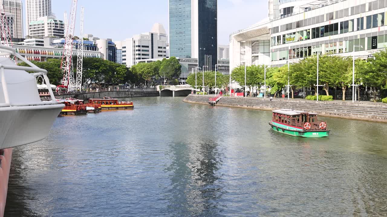 A traditional river cruise boat travels along the Singapore River at Clarke Quay, surrounded by modern buildings and greenery under bright daylight