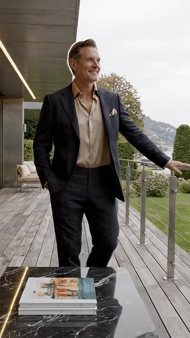 Uomo elegantemente vestito posa con sicurezza su una terrazza moderna che si affaccia su un paesaggio scenografico, emanando fascino e raffinatezza in un abbigliamento elegante.