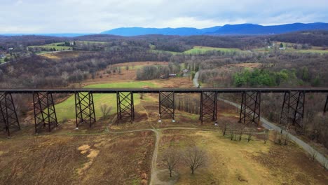 Aerial-drone-video-footage-of-a-train-bridge-viaduct-running-over-a-valley-in-the-Appalachain-Mountains-during-early-spring-on-a-cloud-day,-surrounded-by-mountains-and-farmland