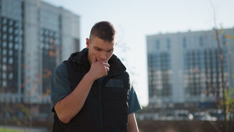 young man in dark vest inhales vape and coughs while holding chest with visible discomfort standing outdoors in urban area with blurred residential high-rise buildings in background