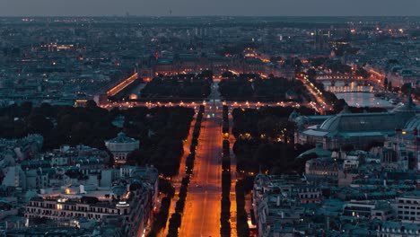 aerial drone footage of champ de mars park and central city of paris during sunset or sunrise with city lights being yellow and orange down below. cars and vehicles driving on road between buildings.