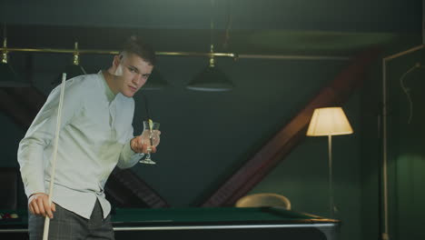 young man in white shirt and checkered trousers holds cue stick and sips lemon drink with black straw while pacing beside green pool table, observing ball arrangement under focused lighting