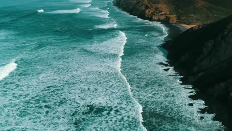 restless surf of blue-turquoise ocean meets cliffs and sandy bay in light of setting sun, drone aerial panorama