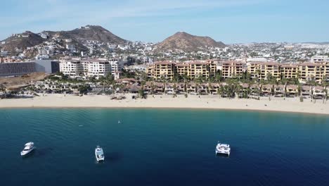 aerial shot over a travelers paradise over the coast in front of medano beach in cabo san lucas, mexico with view of the floating boats, gorgeous beach and hotel complexes with mountain scenery