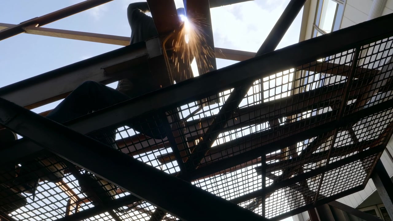 Arbeider som sveiser en stål trappeplattform på et industriområde, med oransje gnister, bjelker, rekkverk, netting og lys himmel synlig fra lav vinkel, som viser fokusert innsats, metallteksturer, struktur, og