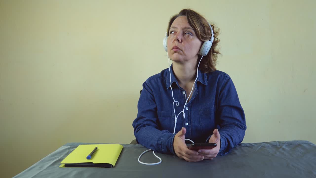 Portrait of a forty-year-old woman in a denim shirt sits at a table in white wired headphones holds a mobile phone. Slow motion portrait