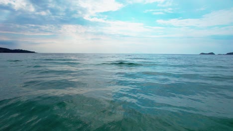 Cámara-Moviéndose-Cerca-De-Las-Olas-Del-Mar-Con-Un-Cielo-Azul-Claro-Y-Una-Pequeña-Isla-En-El-Fondo,-Isla-De-Thassos,-Grecia,-Europa