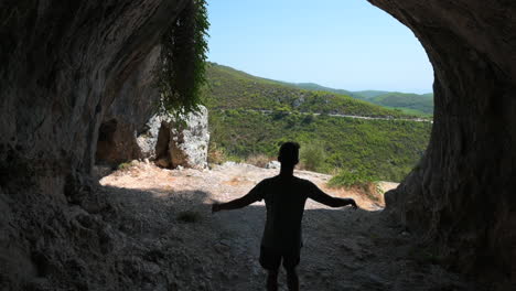 Hombre-Caminando-A-Través-De-Una-Cueva-En-La-Ladera-De-Una-Colina-Con-Una-Vista-Increíble-Del-Valle-Y-Las-Montañas-Circundantes