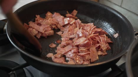 close up on pieces of bacon, toasting in black pan, staring by a wooden spoon 4k