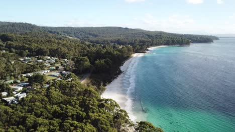 Luftdrohne-Schwenkt-An-Einem-Sonnigen-Tag-über-Einen-Atemberaubenden-Abgelegenen-Strand-Und-Eine-Küstenstadt-Mit-Aquablauem-Wasser-Und-Weißem-Sand,-Umgeben-Von-Grünem-Wald,-Während-Die-Wellen-Hereinrollen