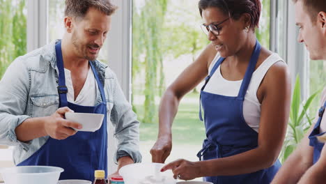 Female-Teacher-With-Male-Student-Mixing-Ingredients-For-Recipe-In-Cookery-Class-In-Kitchen