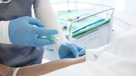 diverse female doctor giving injection to senior female patient in hospital room, slow motion