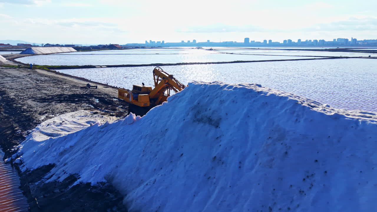 Macchinari pesanti lavorano instancabilmente per modellare cumuli di neve vicino ad acque tranquille, con lo skyline di una città sullo sfondo in una luminosa giornata invernale