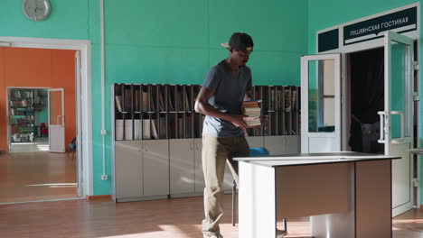 Young man enters building carrying stack of books, crosses room past teal wall cabinets and clock and drops books on desk bathed in sunlight with tidy shelves and minimalist modern decor