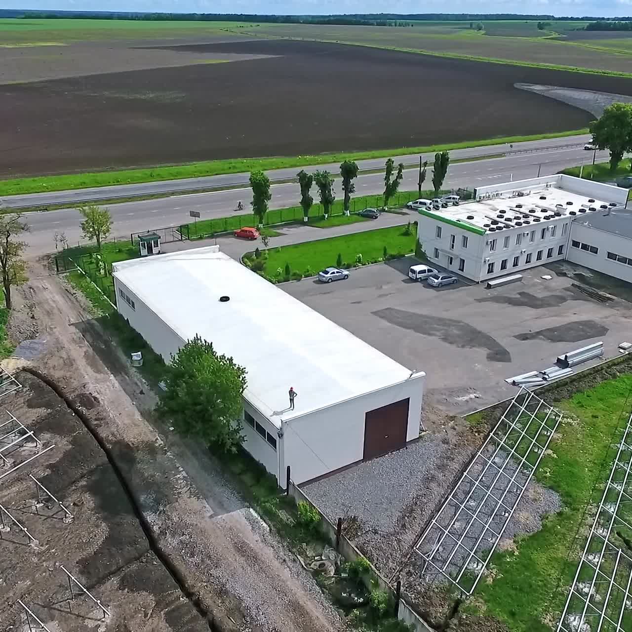 Male worker standing on a roof on the nature background. Engineer in protective helmet on a building among the field with future solar power station. Camera rising up.