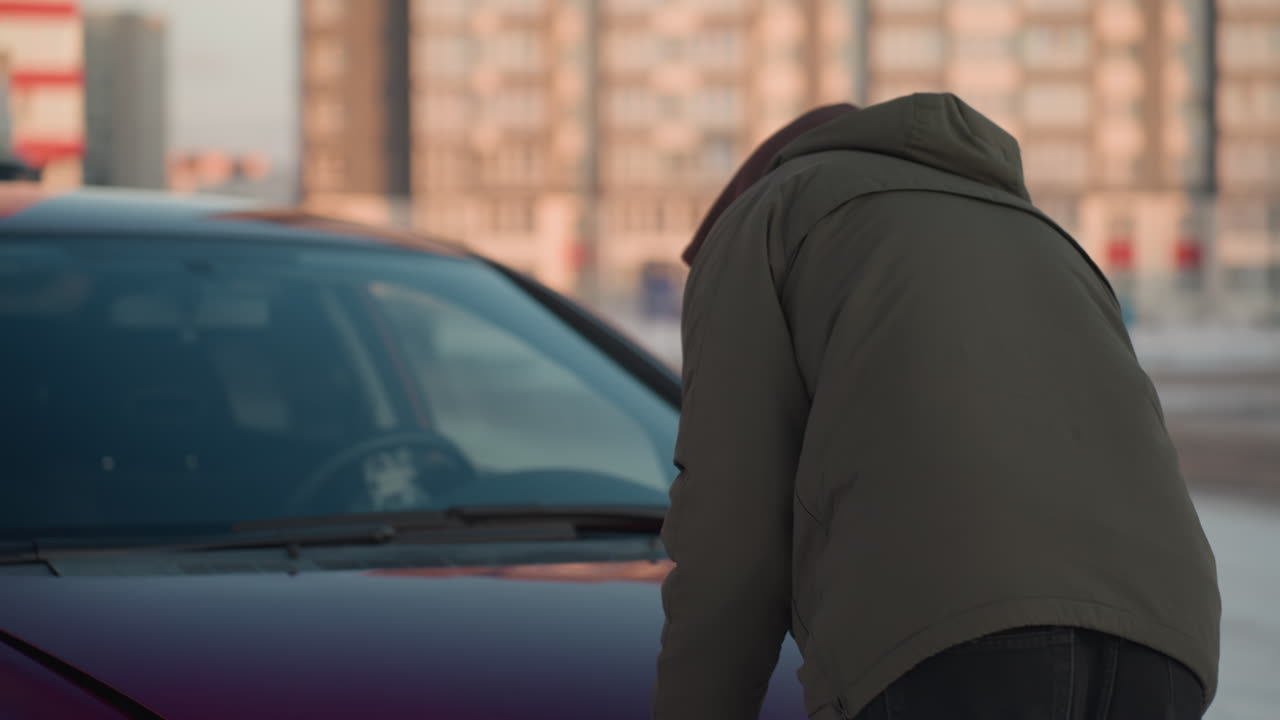 Back view of man in hooded jacket closing car bonnet in cold weather, vehicle partially visible, parked in snowy open lot with blurred urban buildings and structures in the background