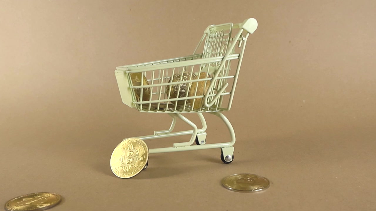 Monete bitcoin fisiche dorate che cadono in un carrello della spesa in miniatura, a simboleggiare l'acquisto e l'investimento in criptovalute, asset digitali e la crescente tendenza dello shopping online