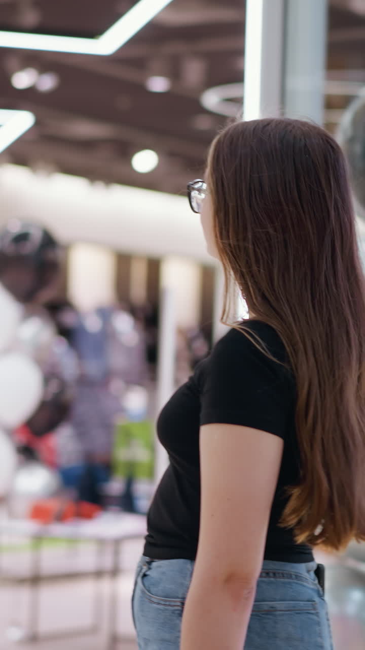 Young lady in glasses walking in shopping mall with bokeh lights in background, surrounded by store displays featuring fashionable clothing, bright lighting, and modern shopping environment