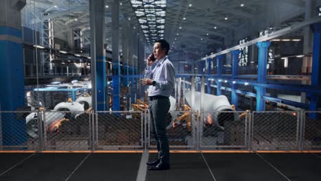 full body side view of an asian male professional worker standing at center infront of the wind turbine factory, he is speaking with someone in the phone and moving his left hand
