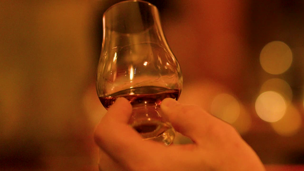 Close-up of hand swirling whisky glass, amber liquid, warm ambient lighting, shallow focus, bar setting