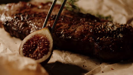 placing fig with kitchen tongs as garnish next to cooked ribeye steak, close up