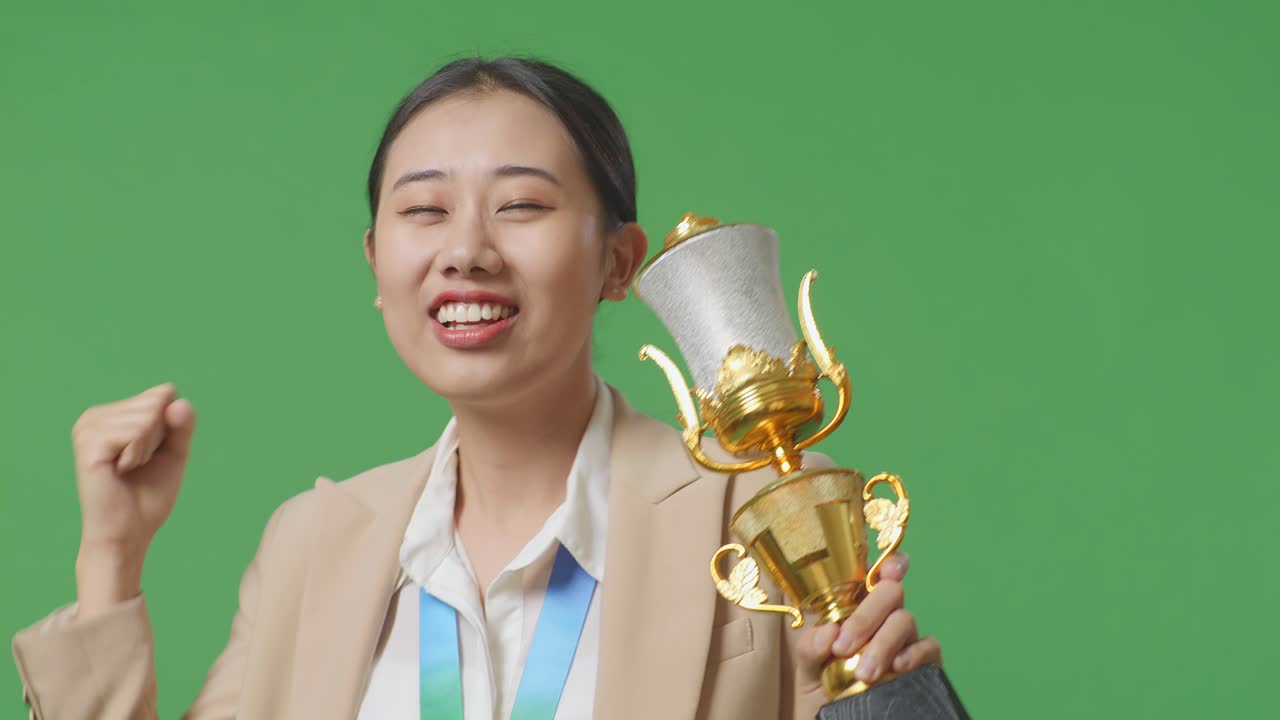 금메달을  슈트를 입은 아시아 사업가 여성이 금 트로피를 바라보고 스튜디오의 녹색 스크린 배경에서 첫 번째 우승자로 우승을 축하합니다.