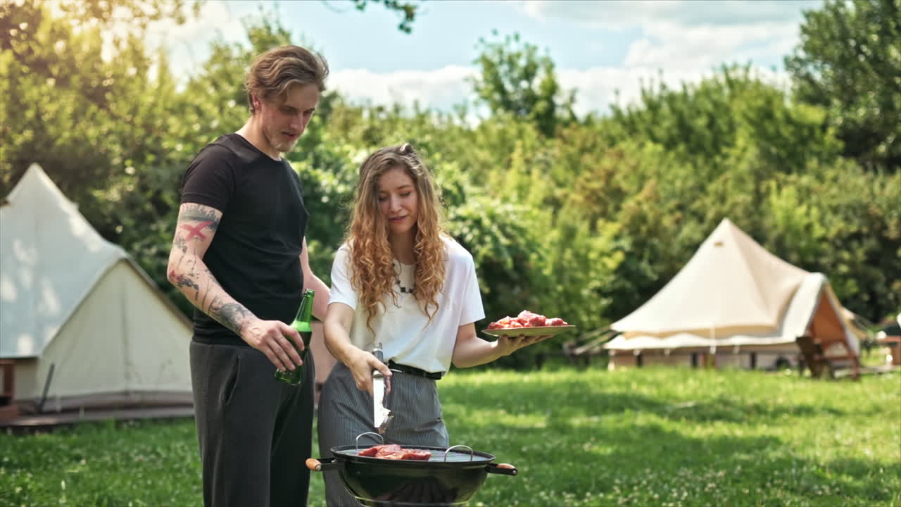 Young couple frying meat on the grill and drinking beer, laughing. Greenery around. Glamping. Slow motion