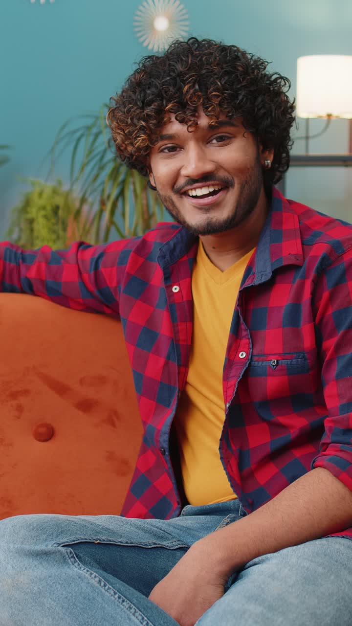 Uomo indiano che sorride amichevolmente alla telecamera, agita le mani, fa un gesto, saluta, saluta a casa sul divano.