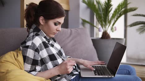 Relaxed-pretty-girl-sitting-comfortably-on-a-couch,-laptop-on-her-legs.-With-a-card-in-her-hand,-typing-something,-searching.-Side-view