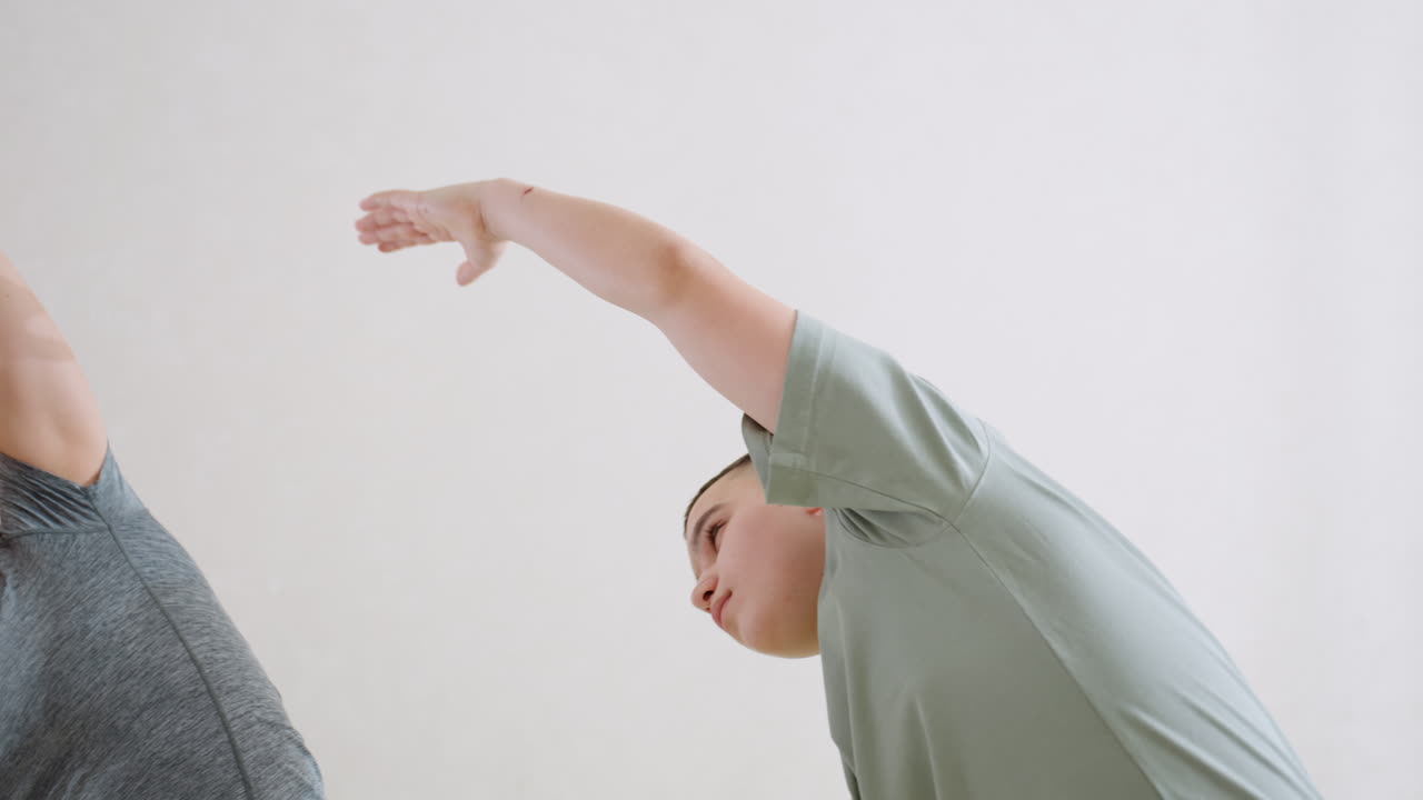 여성이 유연성, 근력, 협응력, 에너지를 키우는 실내 운동 세션에서 옆으로 팔 스트레칭을 하는 동안 균형, 팀워크를 보여주는 지지적인 지침으로 소년을 지도하고 있어요.