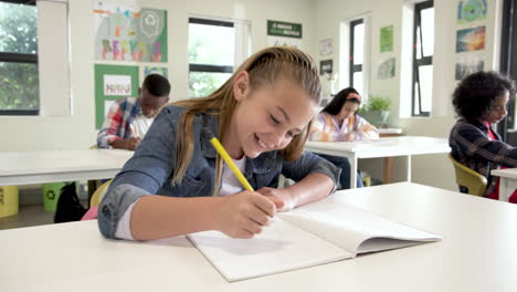 smiling girl writing in notebook in classroom, surrounded by focused classmates, at school