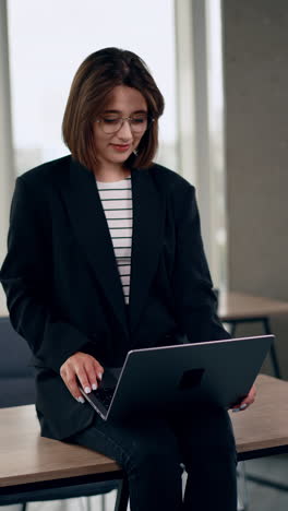 spontaneous caucasian lady sitting on the desk. smiling girl looking at laptop concentrated. vertical video.