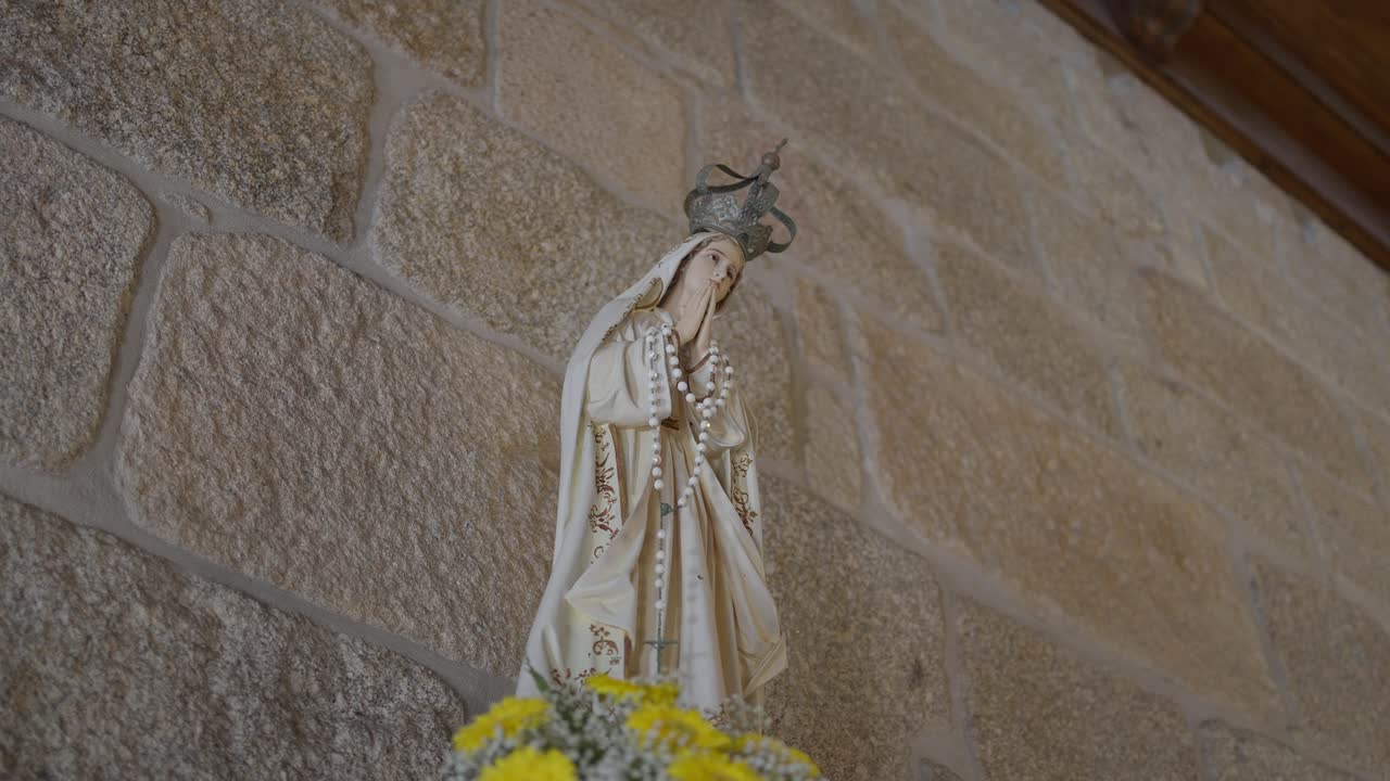 Statue of Virgin Mary with crown, holding a rosary, placed on an altar adorned with yellow flowers, set against a stone wall inside a church