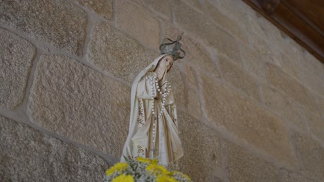 estatua de la virgen maría con corona, sosteniendo un rosario, colocada en un altar adornado con flores amarillas, puesta contra una pared de piedra dentro de una iglesia