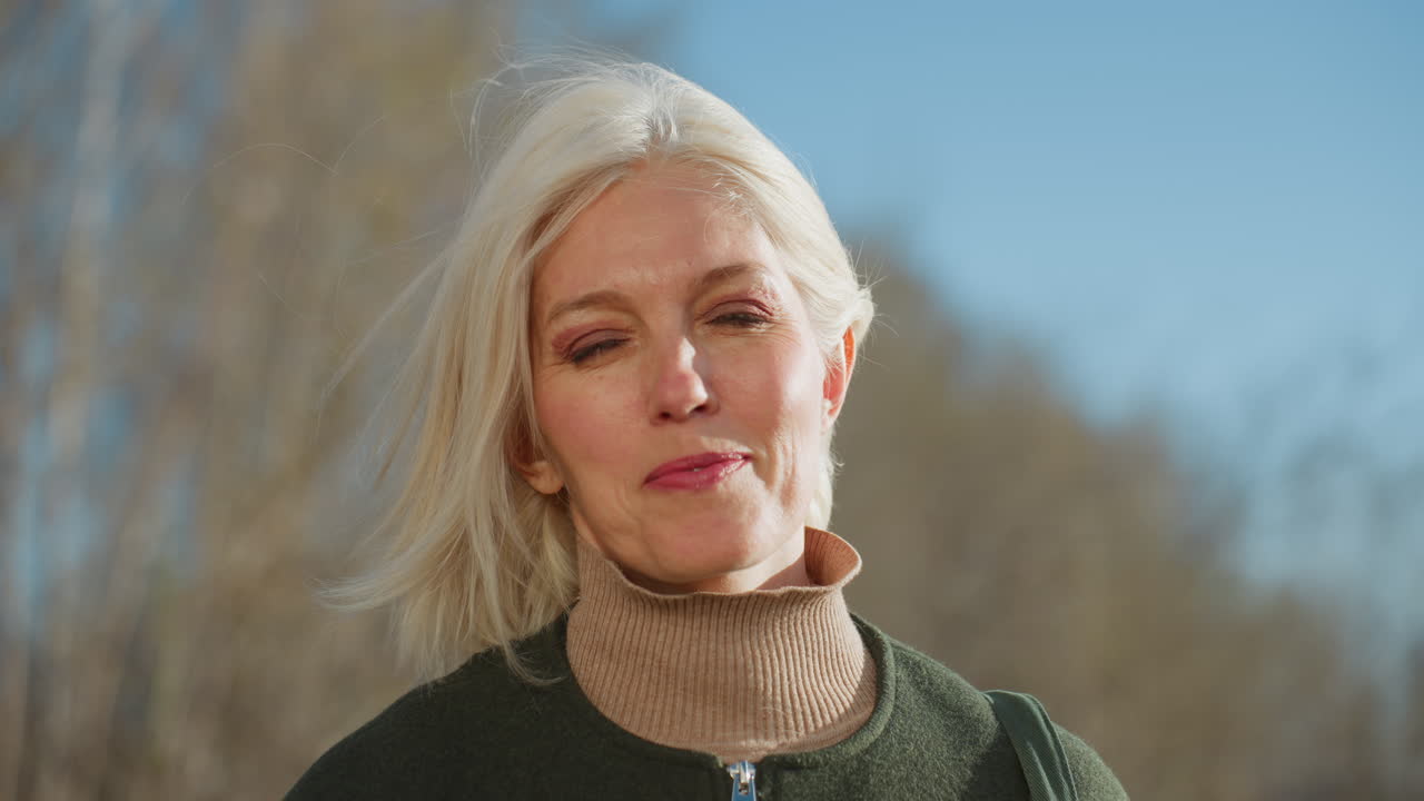 Donna bionda con occhiali da sole, cappotto verde e dolcevita beige, in piedi all'aperto sotto il cielo blu, che guarda in lontananza con espressione sicura, a rappresentare lo stile di vita moderno