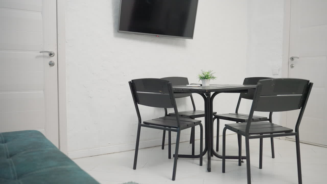 Modern living space with green velvet sofa, black dining table, small potted plant on table, and wall-mounted flat screen TV. Bright white walls and teal curtains enhance the inviting atmosphere
