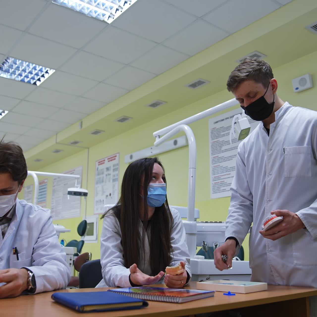 Medical students sit in row at desk and examine models of teeth carefully. Man standing at the desk puts dental instrument to the denture model
