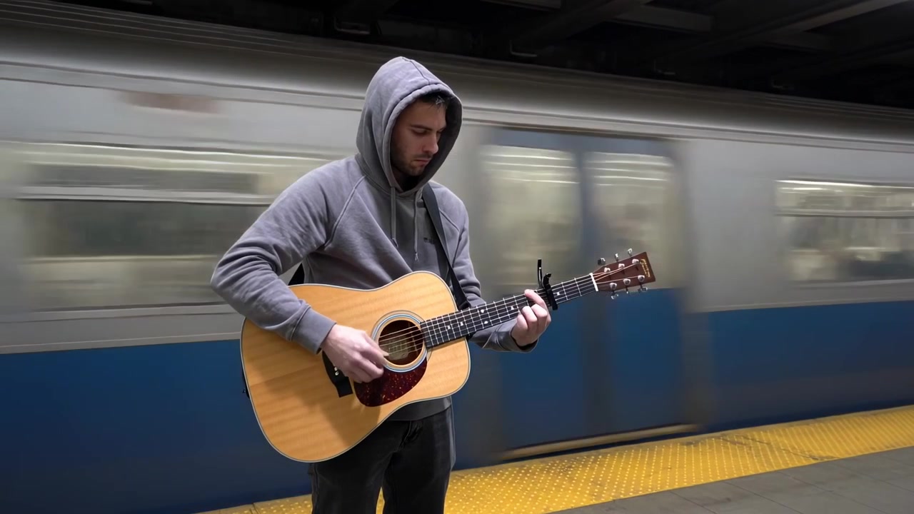 Musicista di strada incappucciato che suona sulla banchina della metropolitana, strimpellando una chitarra acustica con capotasto mentre un treno passa