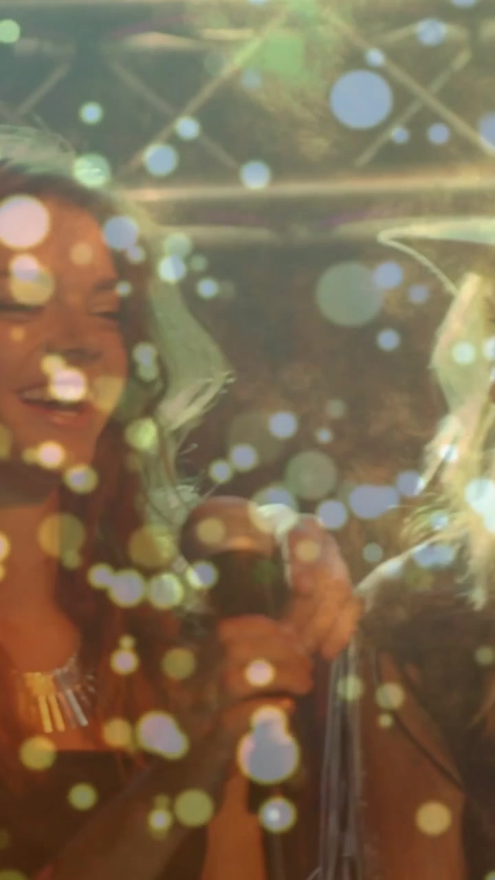 Video verticale: Donna con cappello che alza le braccia mentre il cantante a sinistra porta su il microfono iniziando una canzone, con effetto bokeh.