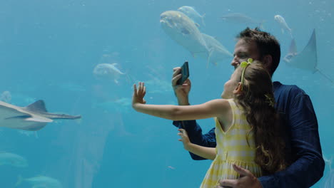 father with little girl at aquarium taking photos of fish using smartphone enjoying marine life swimming in tank having fun at oceanarium sharing on social media