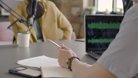 camera focuses on woman hand holding a pen on documents while recording a podcast with unrecognizable man sitting at desk