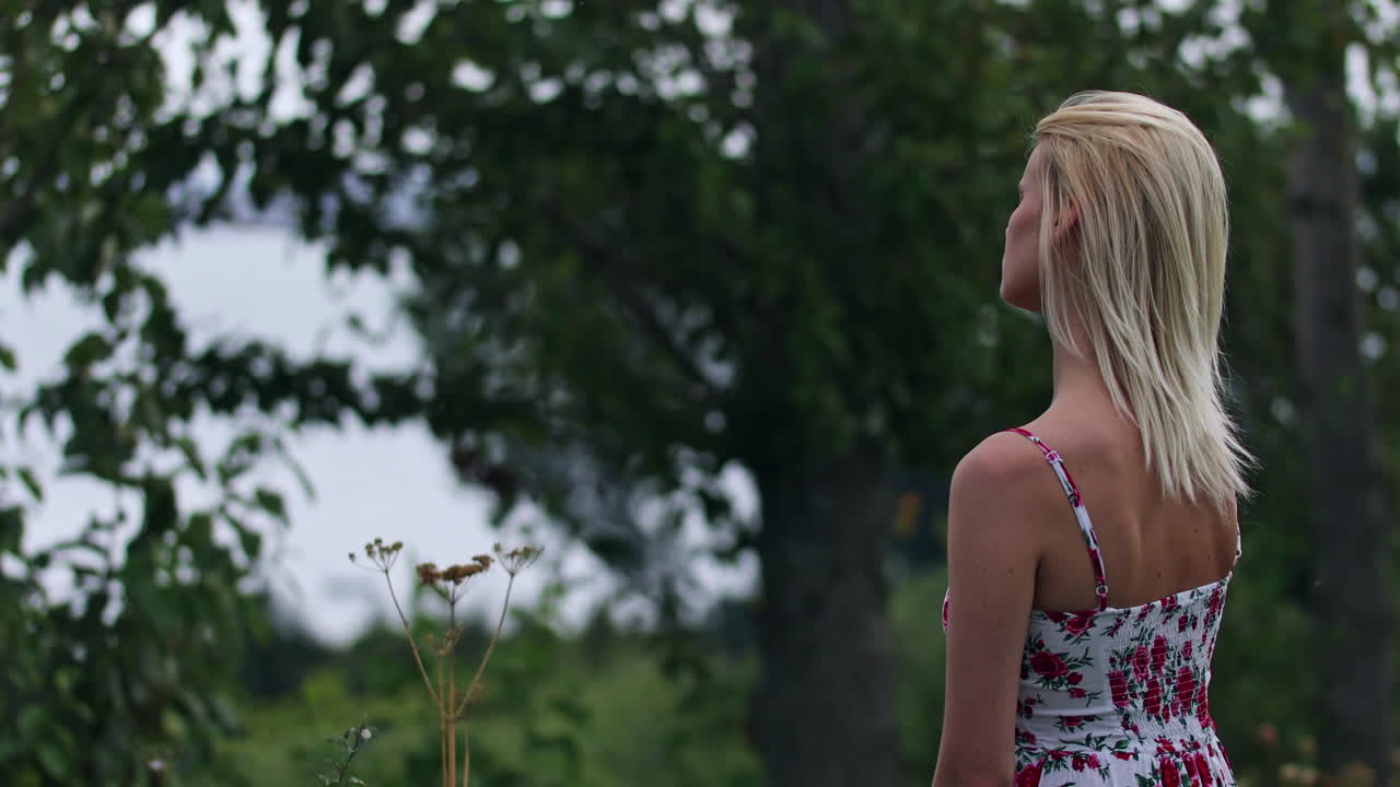 A hipster woman standing alone with a front view reflection of trees. An unhappy lady observing in tranquility with a thoughtful mind. A woman standing in wind weather with an unhappy expression