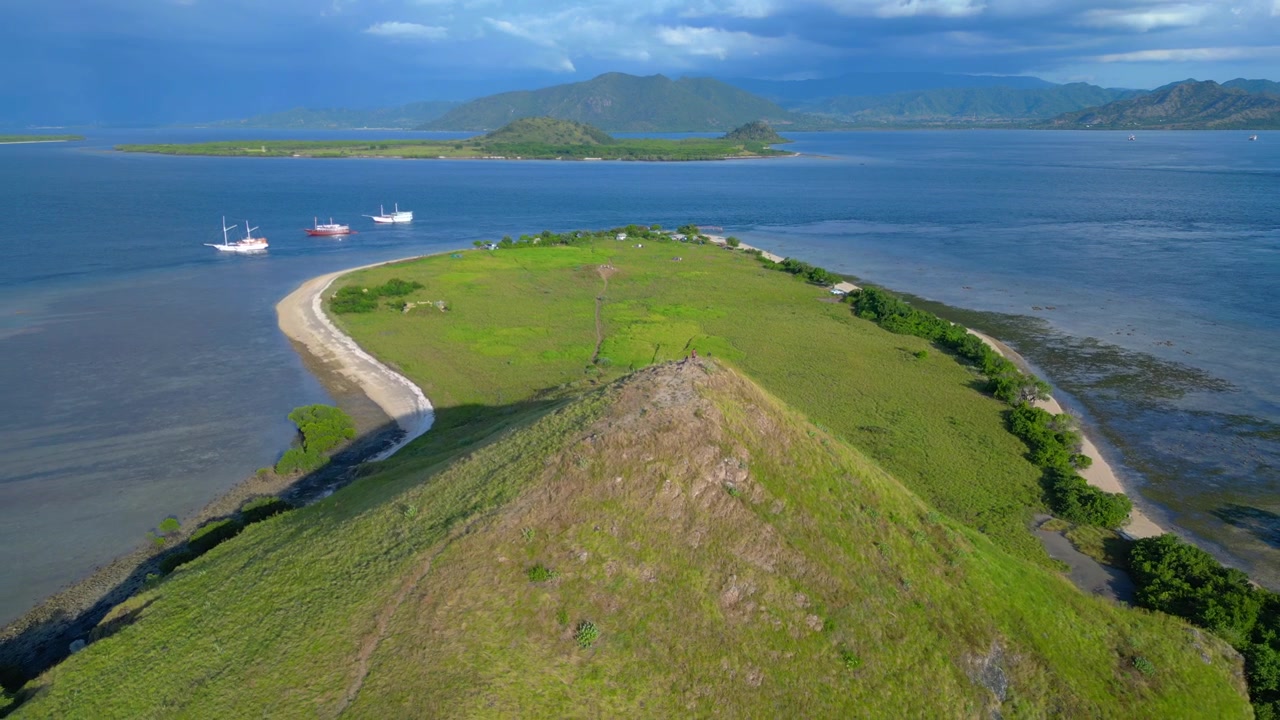 ภูมิทัศน์เกาะโคโมโดพร้อมเนินเขาสีเขียว ชายหาด และเรือประมงพื้นบ้านที่จอดทอดสมออยู่ มุมมองทางอากาศที่น่าอัศจรรย์จากโดรนที่บินแบบพาโนรามาย้อนกลับ